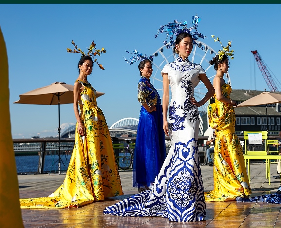 Five figures stand outdoors on a sunny day, wearing elaborate, colorful dresses with long trains in blue, yellow, and white. Each model wears an ornate headpiece. The Seattle Waterfront is visible in the background."